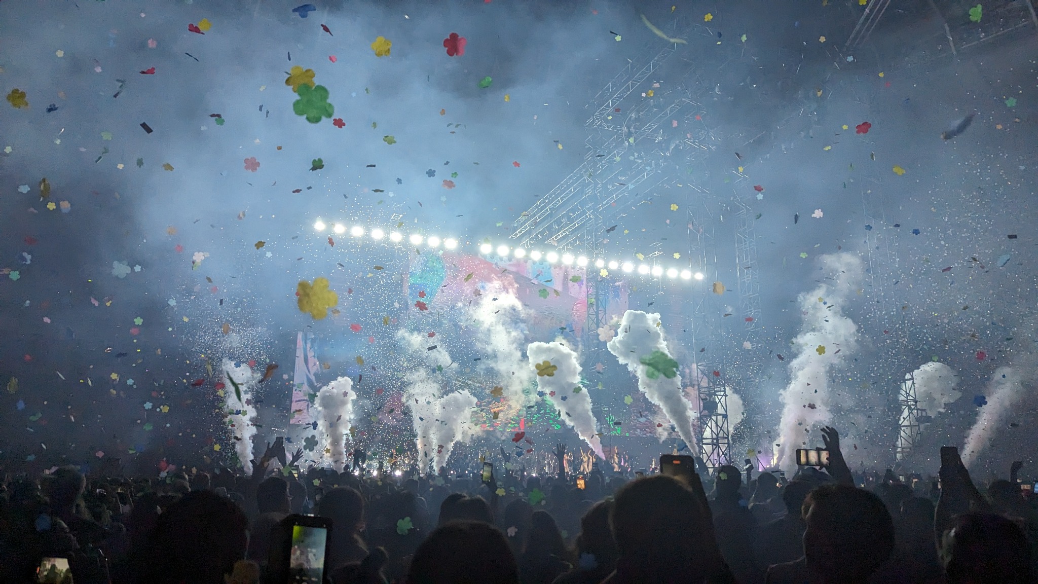 Koncert z tłumem, scena oświetlona reflektorami, konfetti w powietrzu i dym z wytwornic. W tle konstrukcja sceniczna i telebimy. Widok zza publiczności.