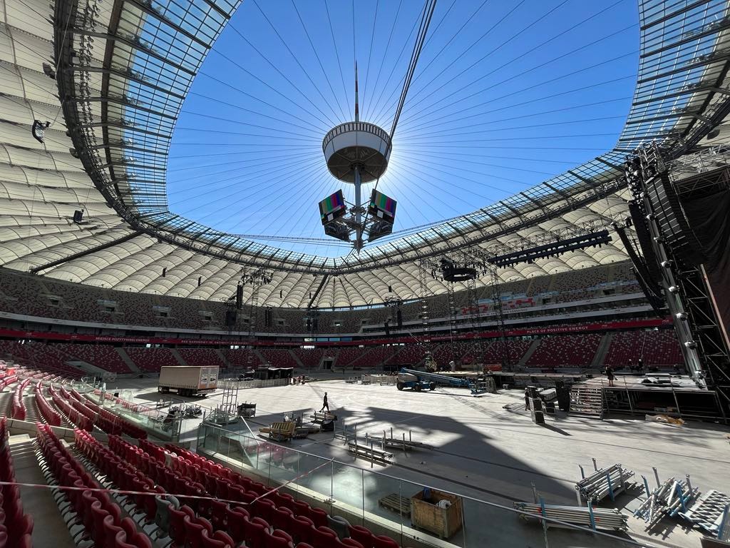 Przygotowania do wydarzenia na stadionie: widoczne konstrukcje sceniczne, oświetlenie i nagłośnienie. W tle charakterystyczna konstrukcja dachu i telebim.