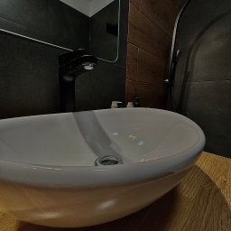 PePe Trade Przemysław Poturalski - Owalna, biała umywalka nablatowa z czarną armaturą na drewnianym blacie, na tle ciemnych płytek i fragmentu kabiny prysznicowej, oświetlona punktowo.