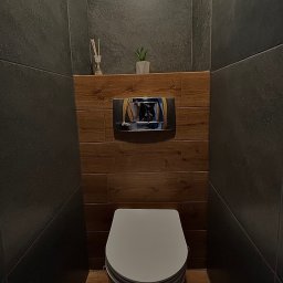 PePe Trade Przemysław Poturalski - Nowoczesna toaleta w ciemnej tonacji z drewnianymi akcentami, widok na wiszącą muszlę klozetową, przycisk spłukujący i dekoracyjne elementy na półce.