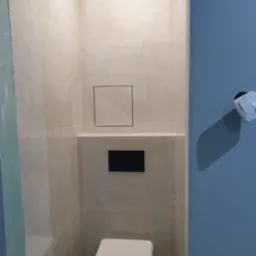 Nowoczesna łazienka z beżowymi płytkami imitującymi marmur, ukryta spłuczka w zabudowie, biała wisząca toaleta i niebieska ściana z włącznikiem światła.