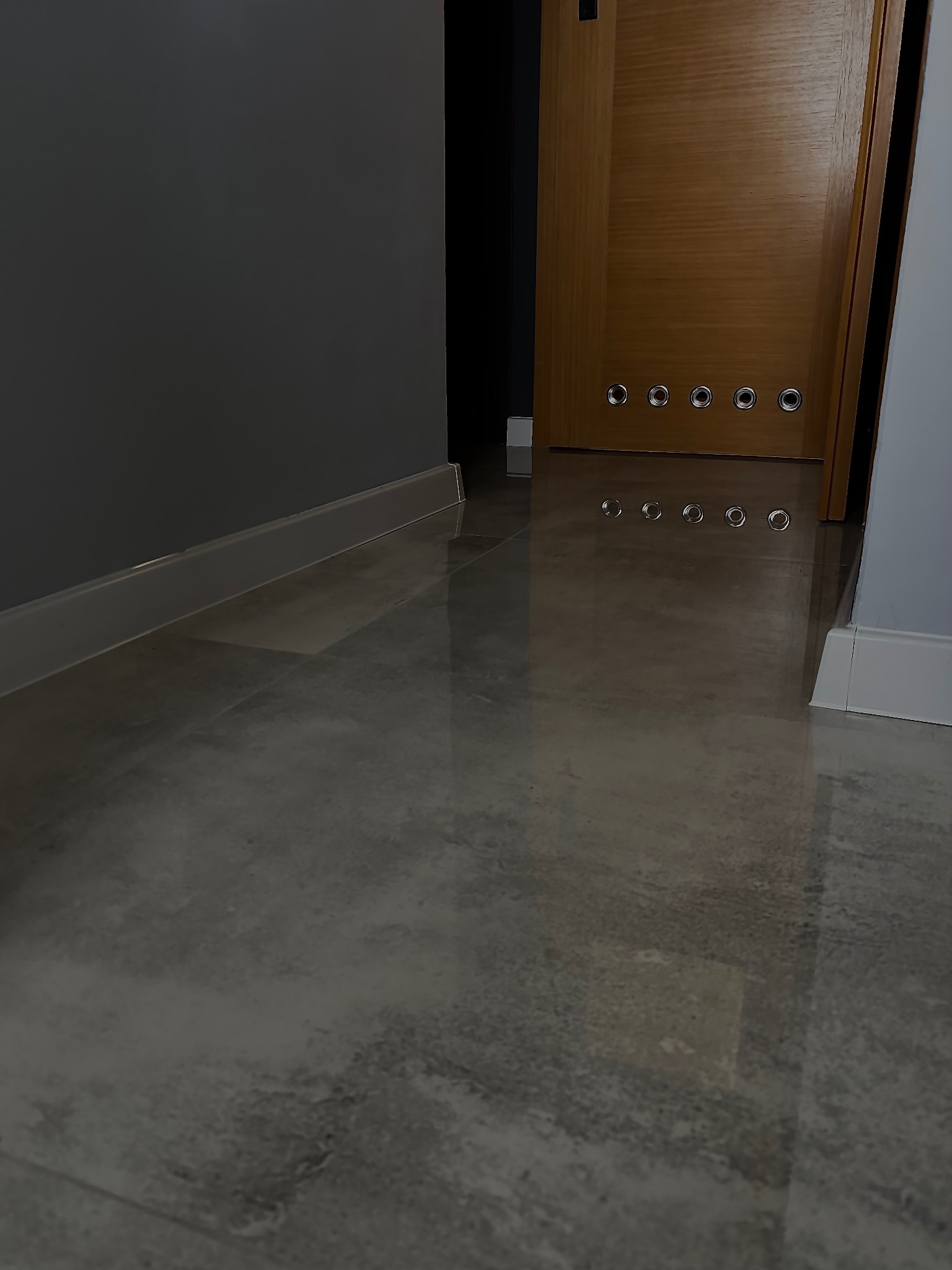 Błyszcząca, szara podłoga z płytek imitujących kamień, widoczna przy podłodze ciemnoszara ściana z listwą przypodłogową oraz fragment drewnianych drzwi z metalowymi oczkami w dolnej części.