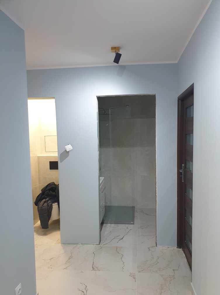 Wnętrze mieszkania w trakcie prac wykończeniowych: widoczne świeżo pomalowane na jasny niebieski kolor ściany, częściowo wykończona łazienka z nowymi płytkami i drzwiami z przeszkleniami,...