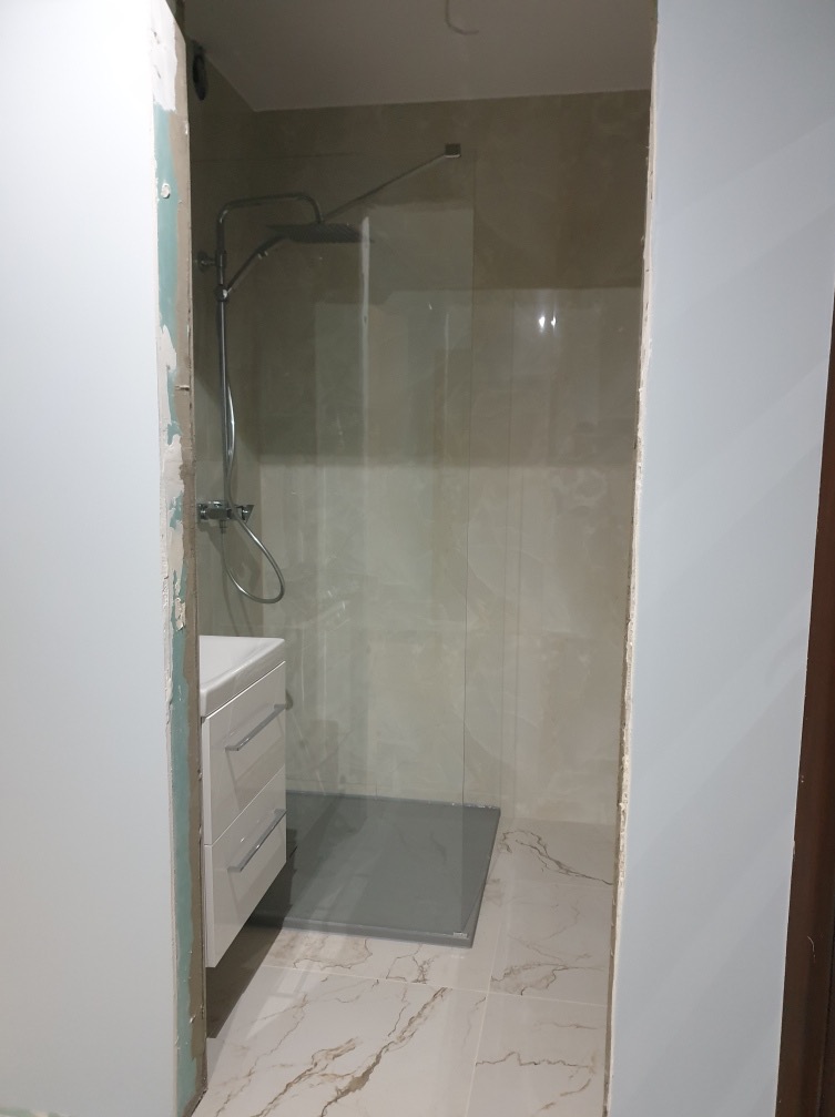 Nowoczesna kabina prysznicowa typu walk-in z szarą brodzikiem i armaturą w kolorze chromu, umywalka z szafką w białym połysku, marmurowe płytki podłogowe, widoczne ślady po remoncie na ścianie.