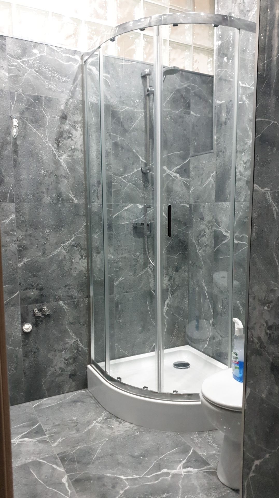 Szara łazienka z kabiną prysznicową narożną, płytki imitujące marmur, widoczny fragment toalety i butelka płynu do czyszczenia.