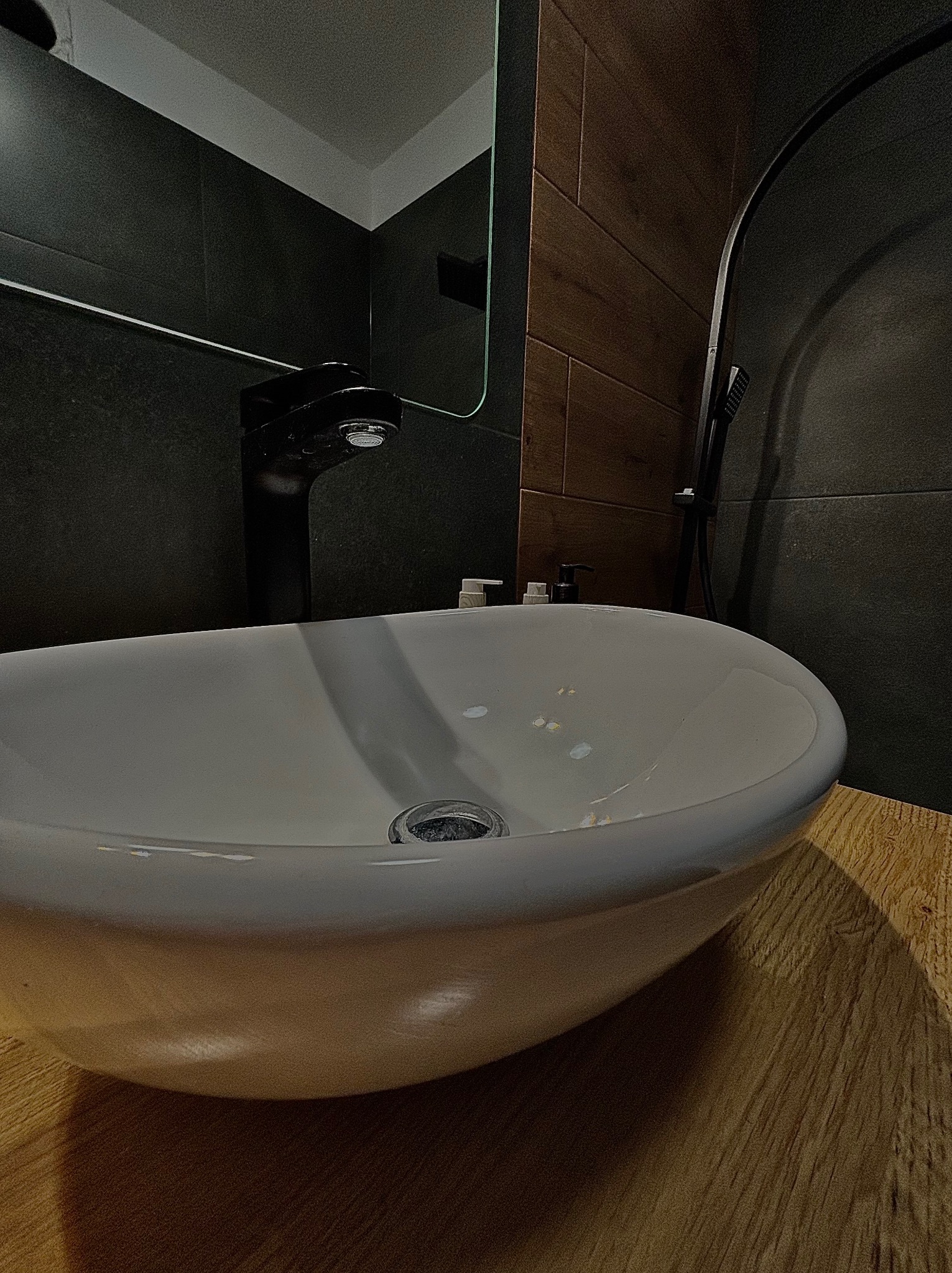 Owalna, biała umywalka nablatowa z czarną armaturą na drewnianym blacie, na tle ciemnych płytek i fragmentu kabiny prysznicowej, oświetlona punktowo.