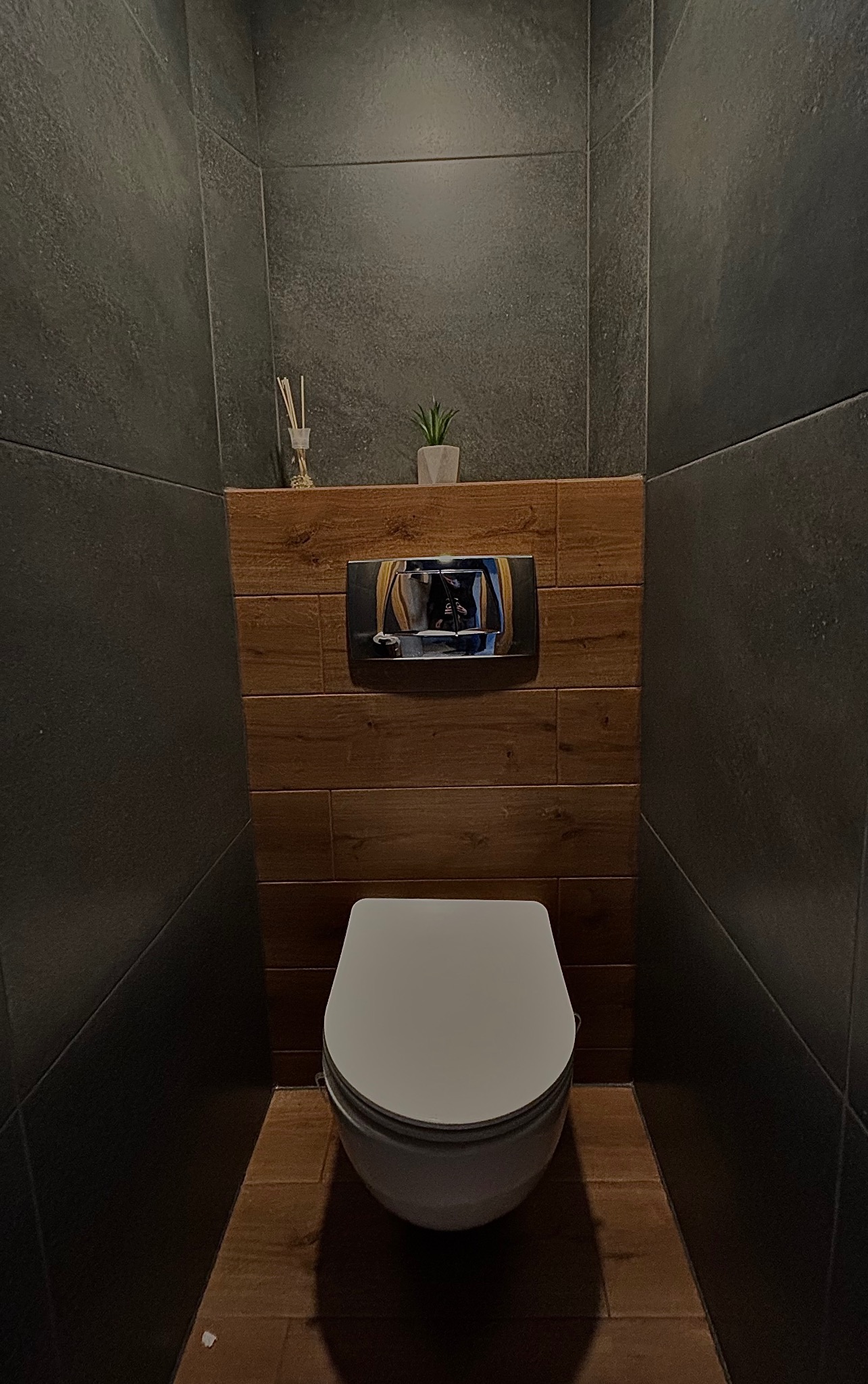 Nowoczesna toaleta w ciemnej tonacji z drewnianymi akcentami, widok na wiszącą muszlę klozetową, przycisk spłukujący i dekoracyjne elementy na półce.