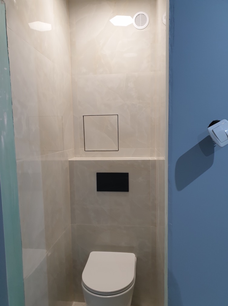 Nowoczesna łazienka z beżowymi płytkami imitującymi marmur, ukryta spłuczka w zabudowie, biała wisząca toaleta i niebieska ściana z włącznikiem światła.