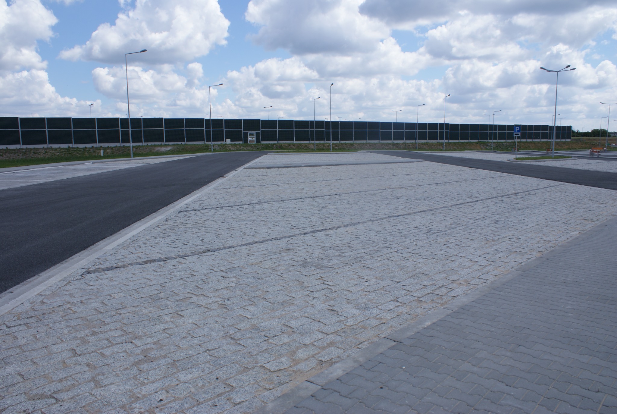 Ukończony parking z kostki granitowej i asfaltu, oddzielony krawężnikiem, z widocznymi miejscami parkingowymi i barierą akustyczną w tle, oświetlony lampami ulicznymi.
