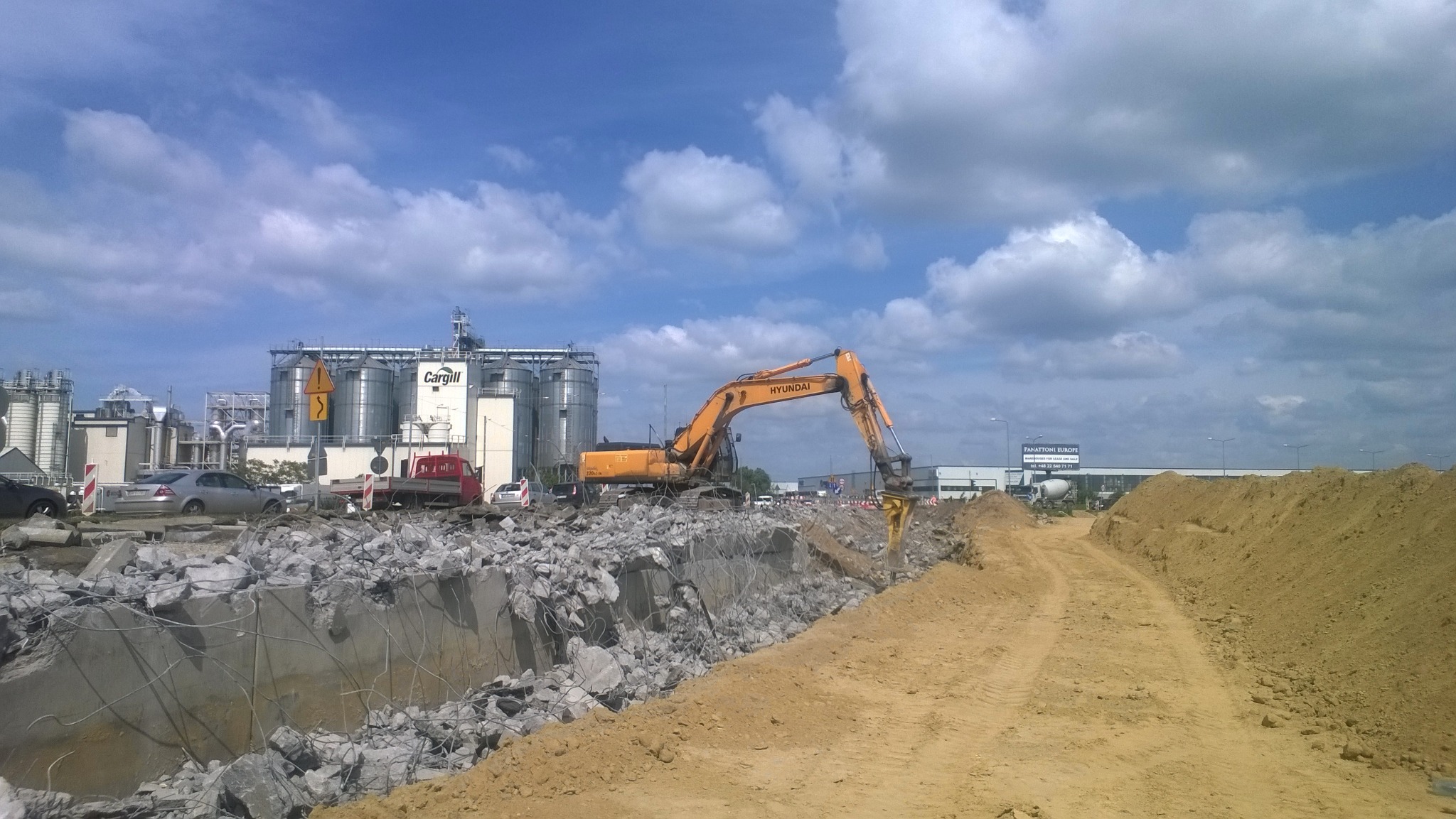 Żółta koparka Hyundai z młotem hydraulicznym rozbija betonową konstrukcję na tle przemysłowego krajobrazu z silosami Cargill i błękitnym niebem.
