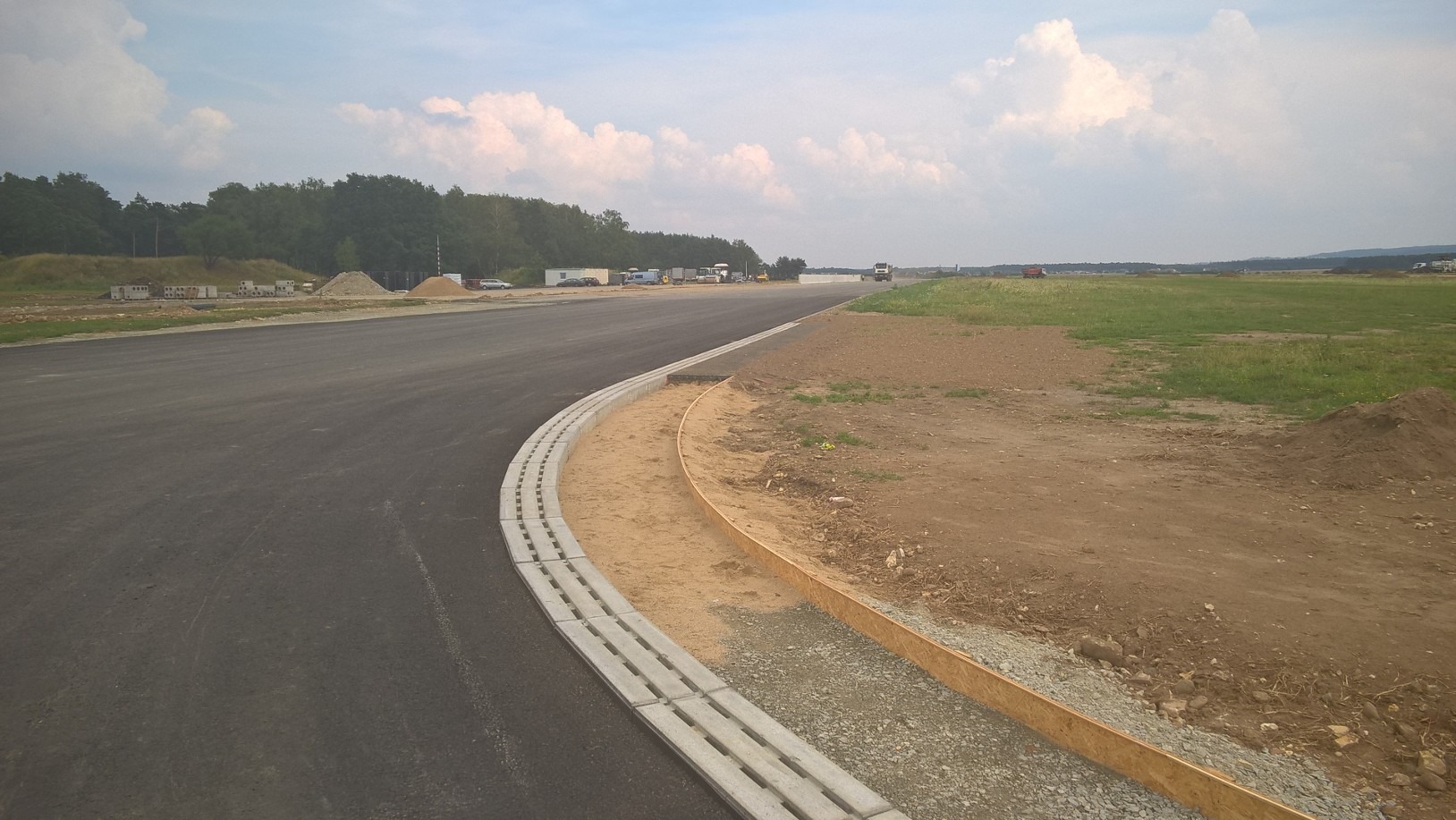 Nowa, gładka nawierzchnia drogi z betonowym korytkiem odwadniającym i przygotowanym podłożem obok, w tle widoczne maszyny budowlane i teren przygotowany pod budowę.