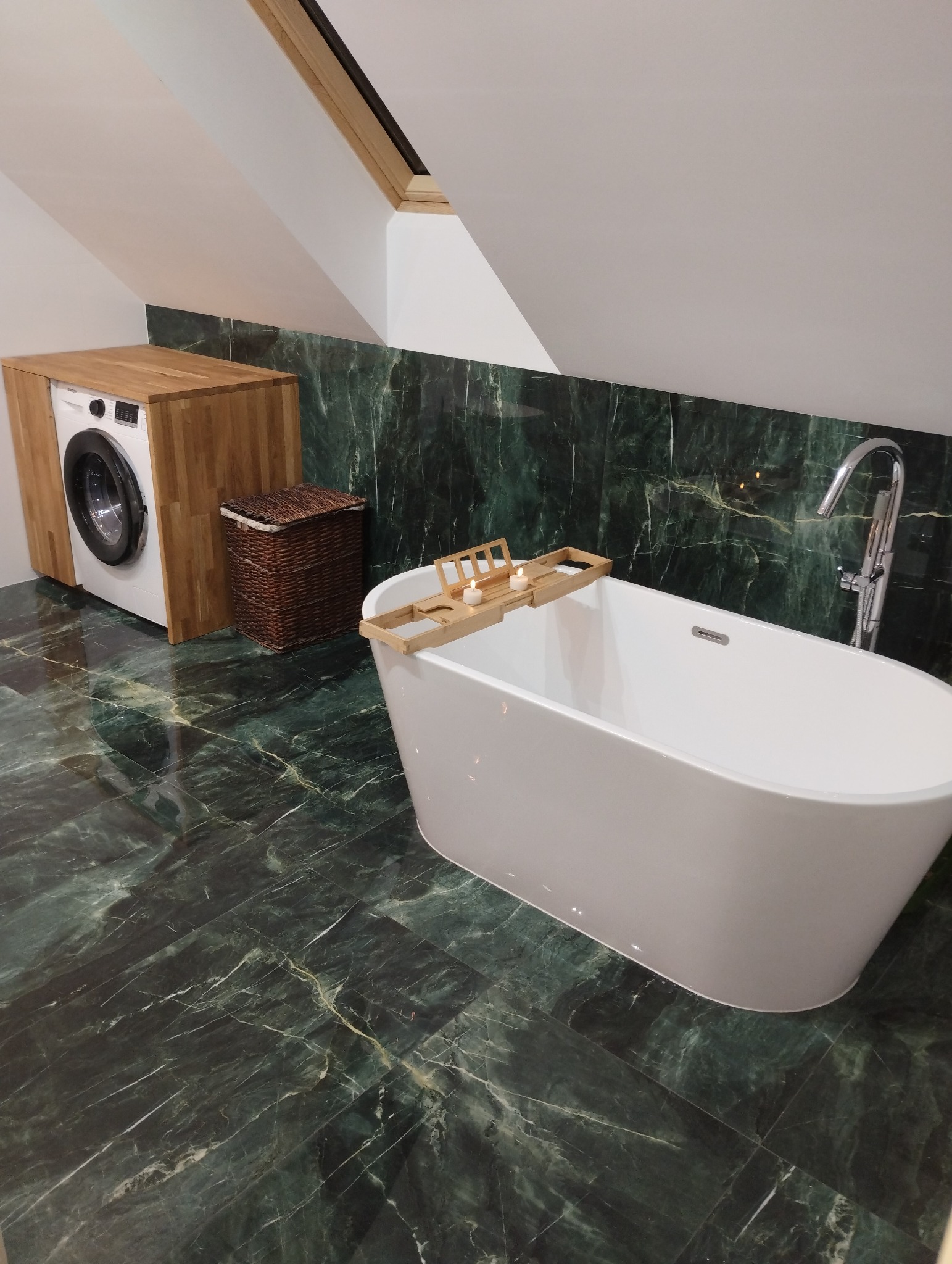 Nowoczesna łazienka na poddaszu z zielonymi marmurowymi płytkami, wolnostojącą białą wanną z drewnianą półką na świece, pralką w drewnianej obudowie i koszem na pranie.