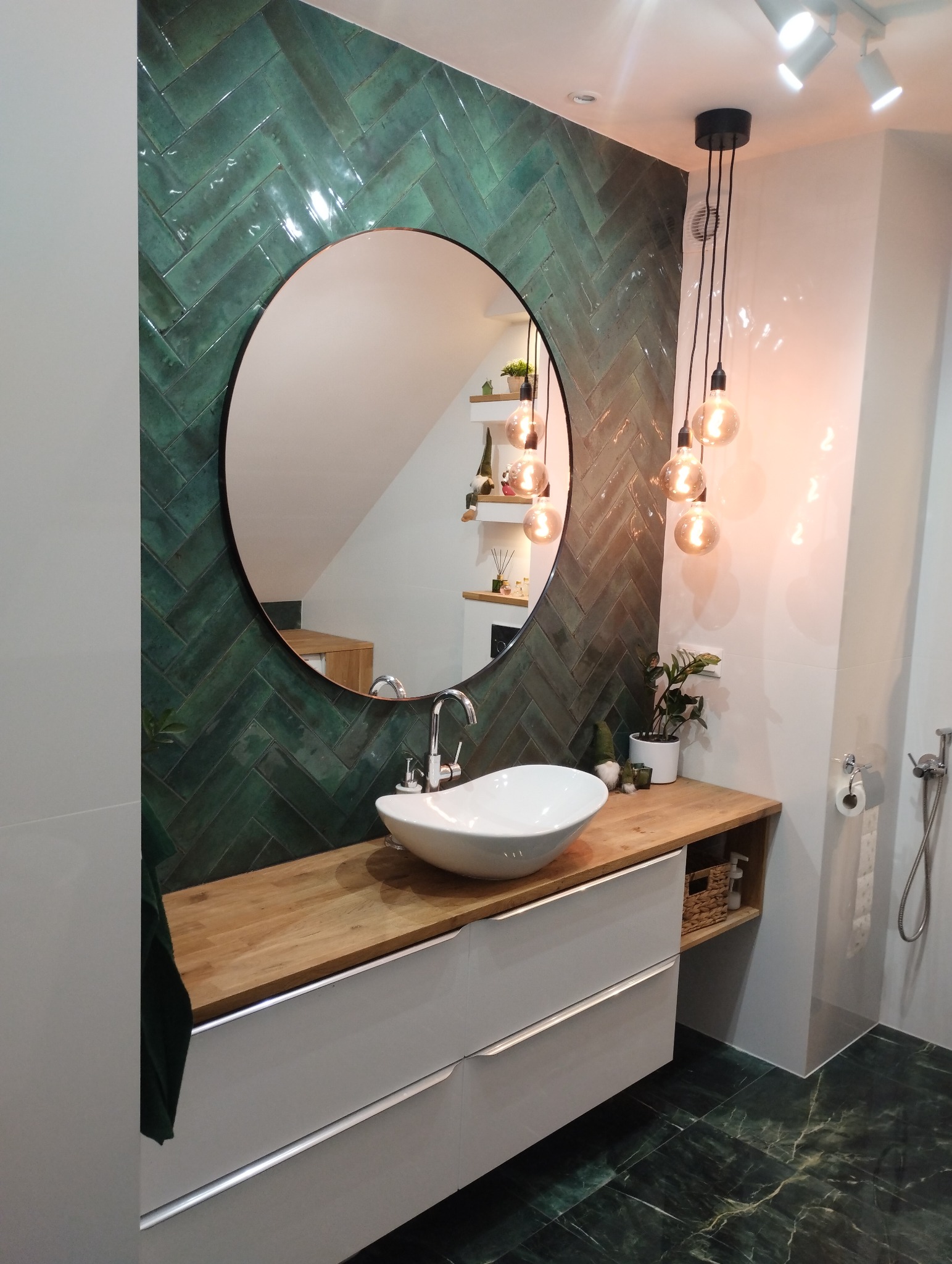 Nowoczesna łazienka w Bydgoszczy z umywalką nablatową, zielonymi płytkami ułożonymi w jodełkę, okrągłym lustrem i wiszącymi lampami z żarówkami Edisona.
