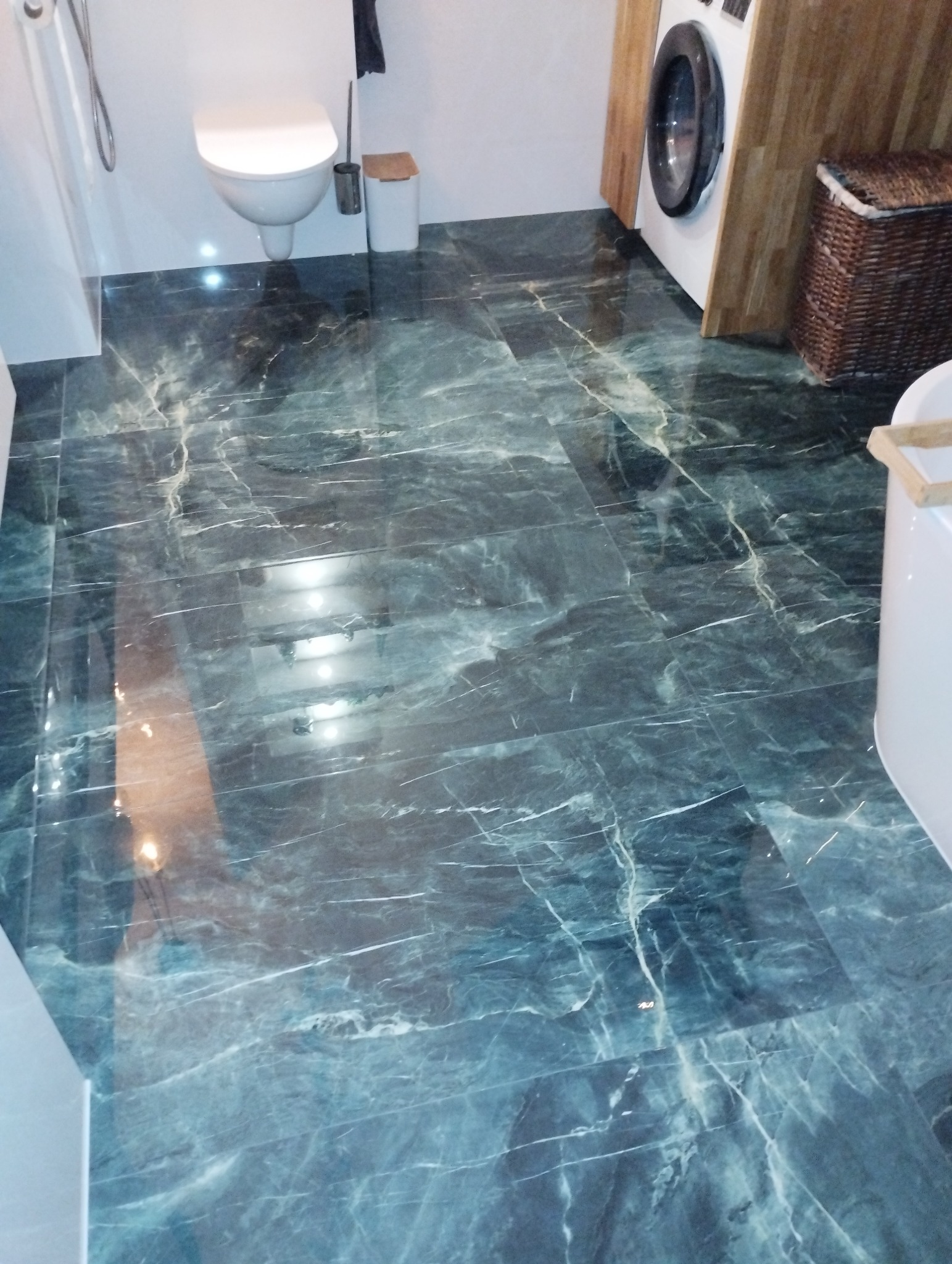 Błyszcząca, ciemnozielona podłoga z płytek imitujących marmur w łazience z toaletą, pralką zabudowaną w drewnianej obudowie i koszem wiklinowym.