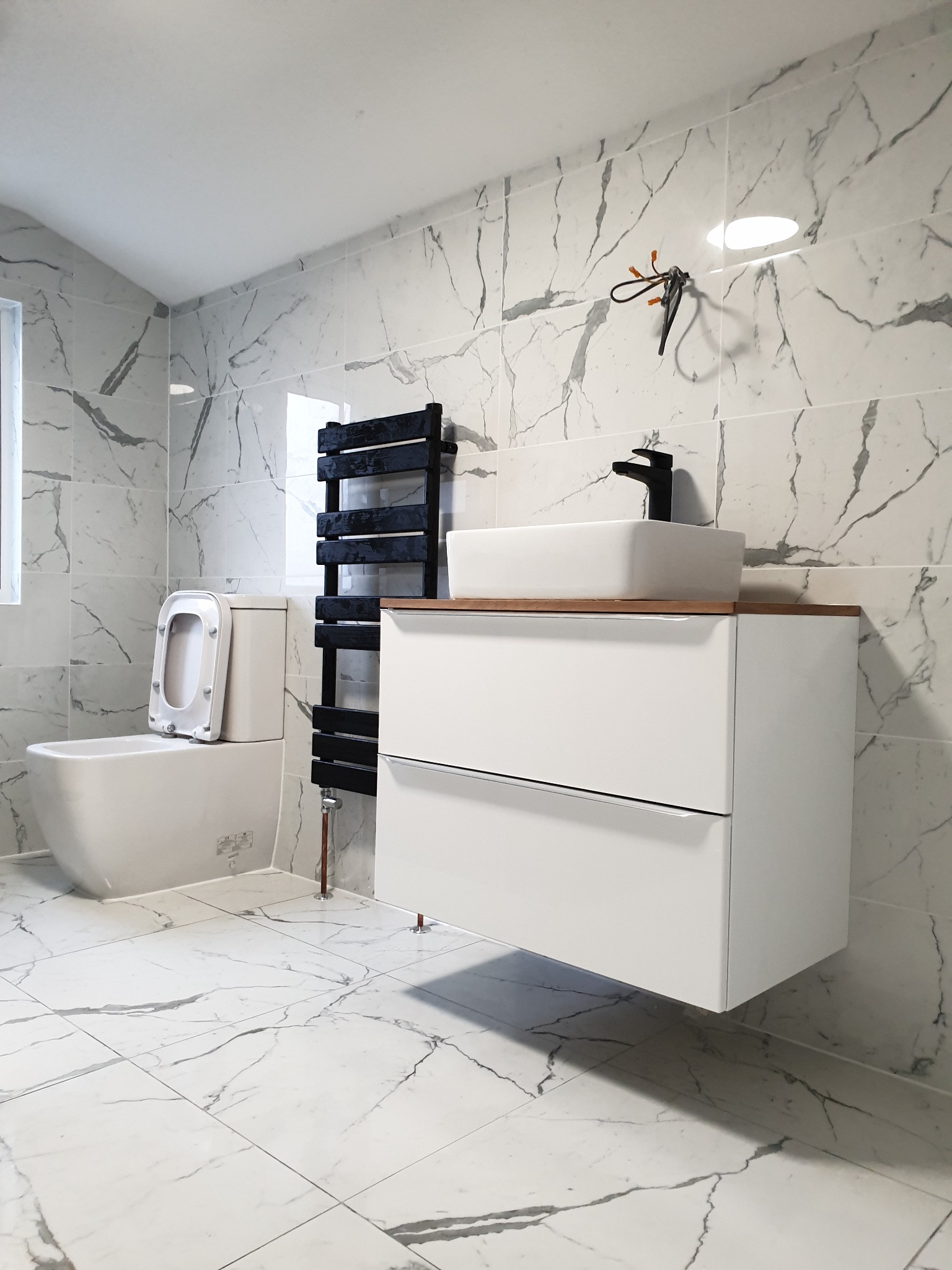 Nowoczesna łazienka z białą armaturą i marmurowymi płytkami, wisząca szafka pod umywalką z drewnianym blatem, czarny grzejnik drabinkowy i toaleta z otwartą deską.