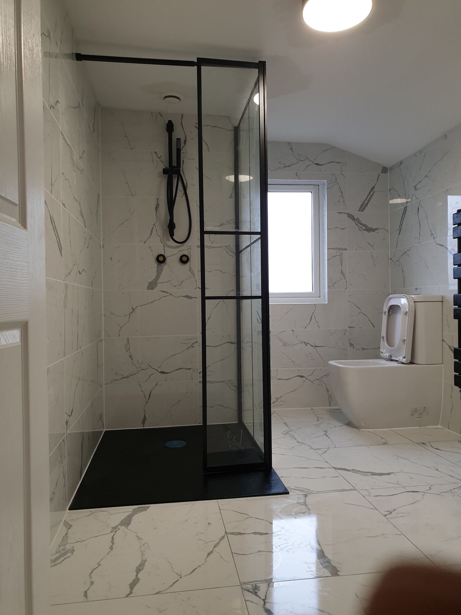 Nowoczesna łazienka z kabiną prysznicową typu walk-in z czarnymi profilami, białe płytki imitujące marmur, wisząca toaleta z podniesioną deską, czarna armatura i grzejnik.