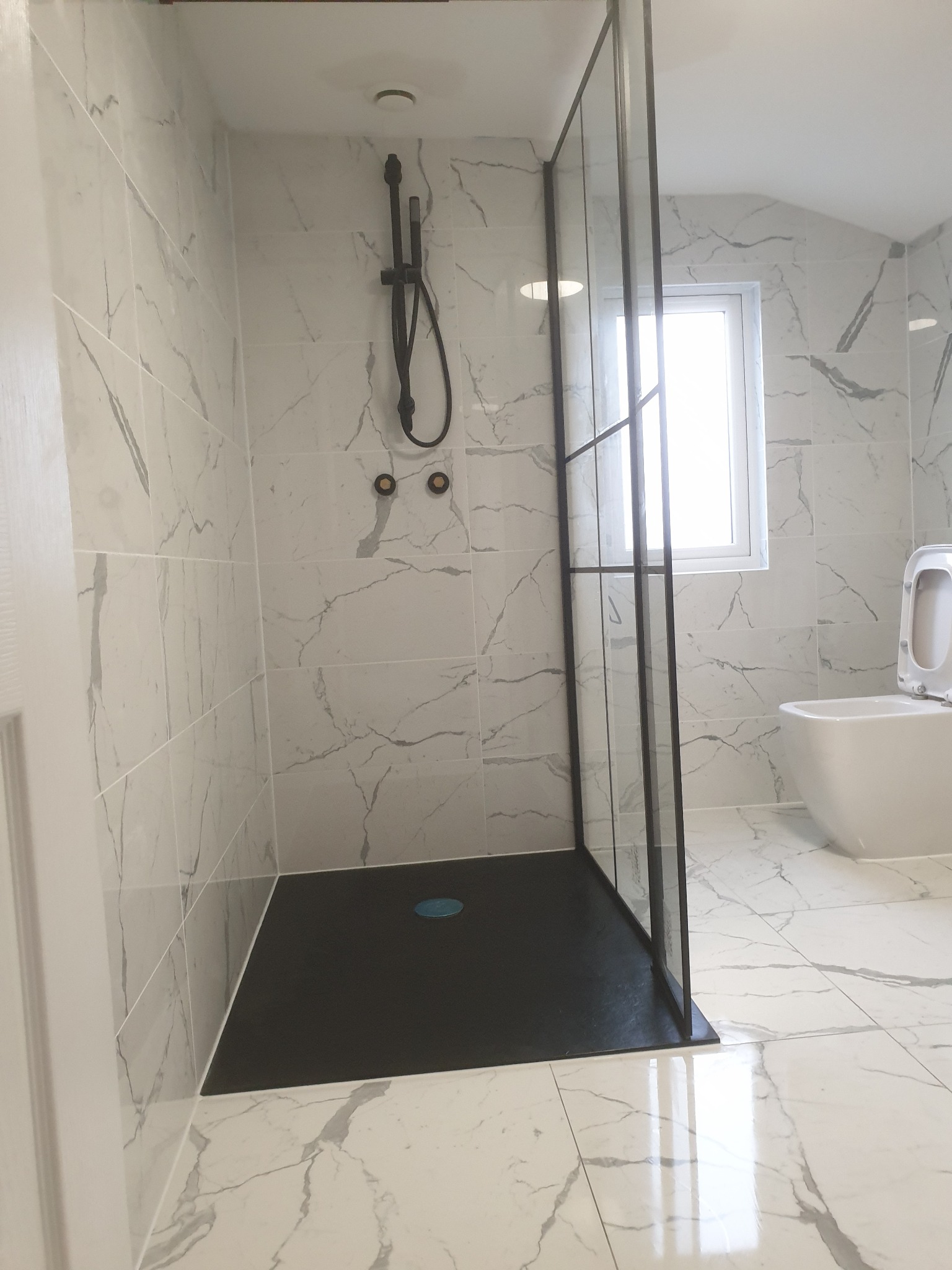 Nowoczesna łazienka z kabiną prysznicową typu walk-in z czarną armaturą i brodzikiem, wykończona białymi płytkami imitującymi marmur, z widoczną toaletą w tle.