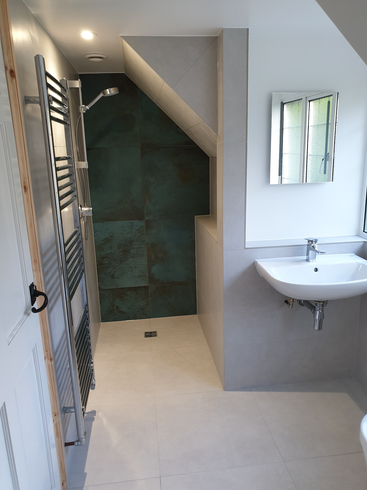 Nowoczesna łazienka z kabiną prysznicową wykończoną w ciemnej zieleni, jasnymi płytkami podłogowymi i umywalką nablatową pod oknem.