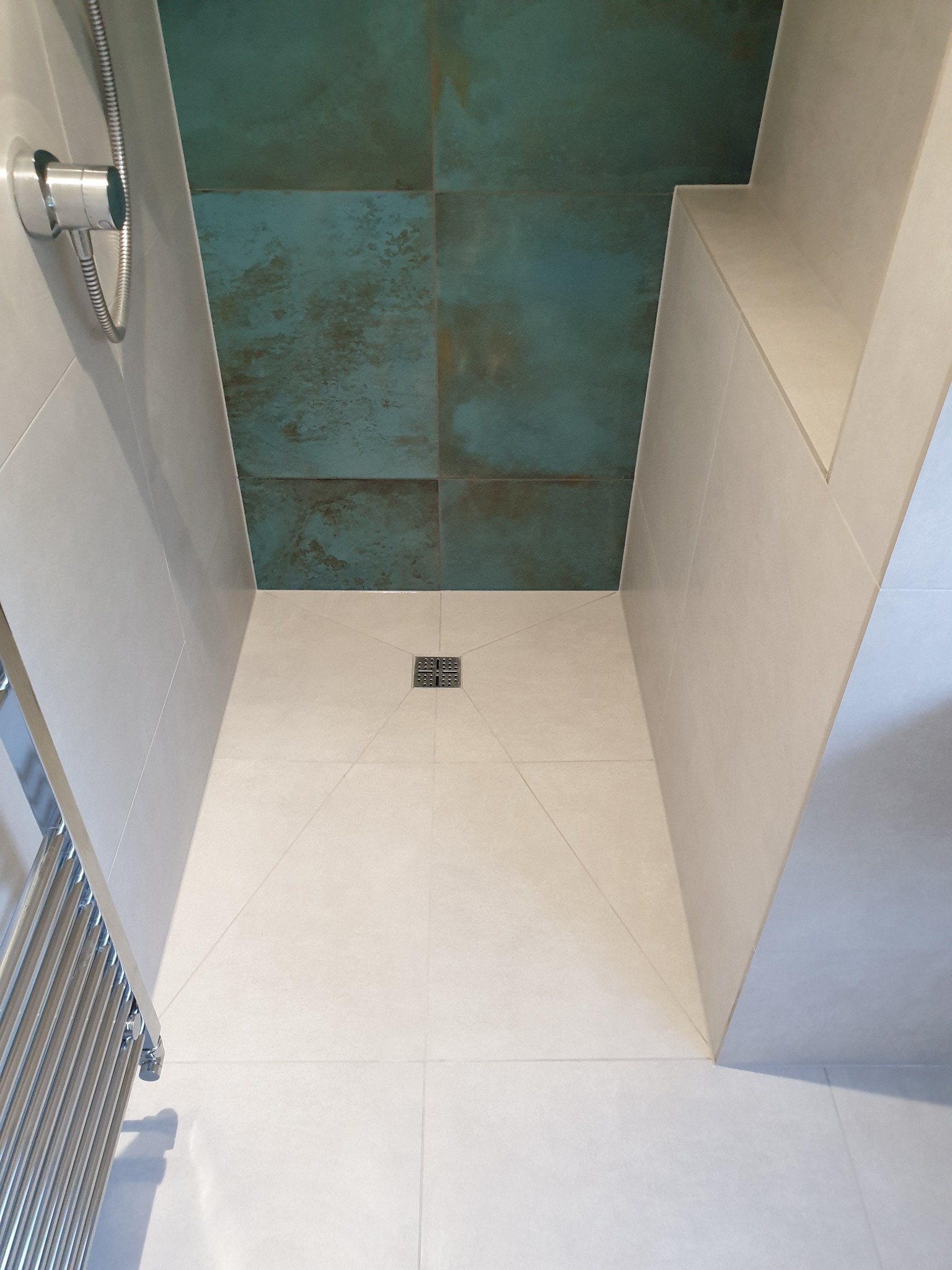 Nowoczesny brodzik bezprogowy wyłożony jasnymi płytkami, z odpływem liniowym i akcentem w postaci ciemnozielonych płytek na ścianie, widoczny grzejnik drabinkowy.