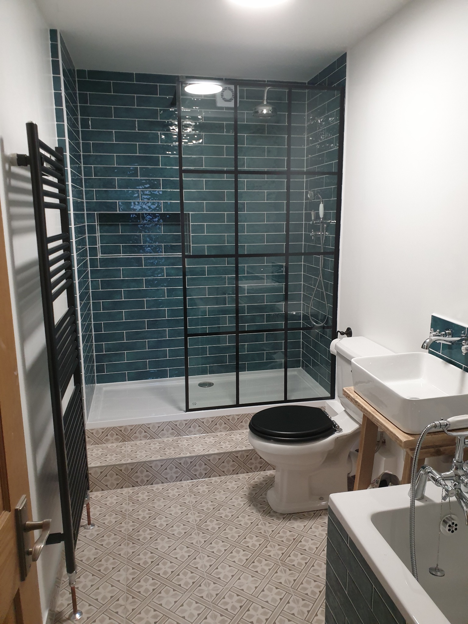 Nowoczesna łazienka z kabiną prysznicową typu walk-in z czarnymi szprosami, turkusowe płytki na ścianach, biała ceramika łazienkowa i grzejnik drabinkowy w kolorze czarnym.