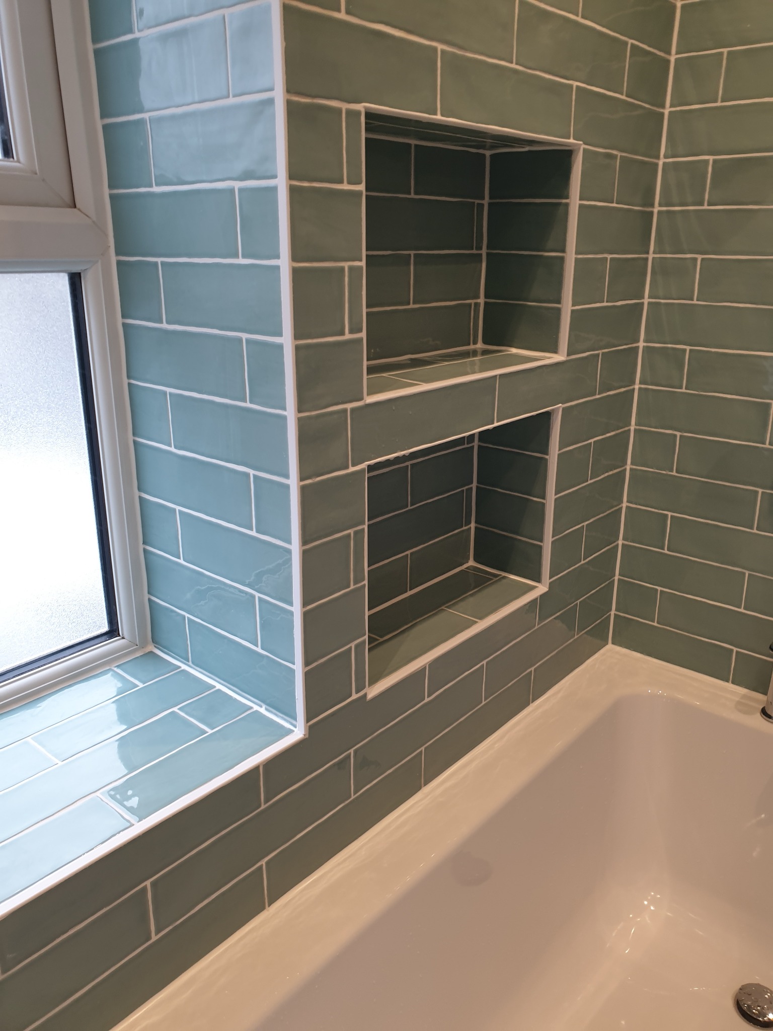 Wykończona łazienka z turkusowymi, prostokątnymi płytkami ułożonymi w cegiełkę, wbudowane półki w ścianie obok okna i krawędzi wanny.