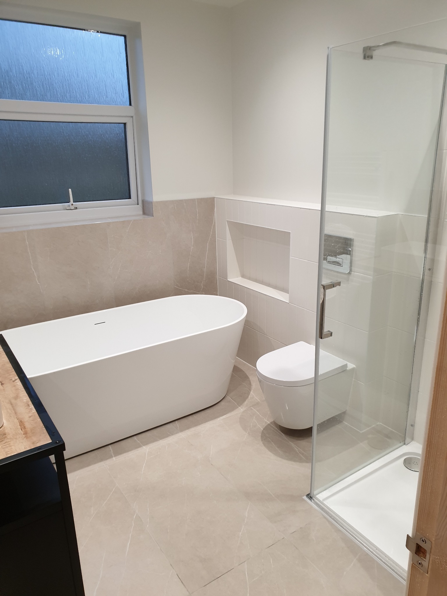 Nowoczesna łazienka w jasnych barwach: wolnostojąca wanna, podwieszana toaleta i przeszklona kabina prysznicowa z chromowanymi detalami, wykończona beżowymi płytkami imitującymi marmur, oświetlona...