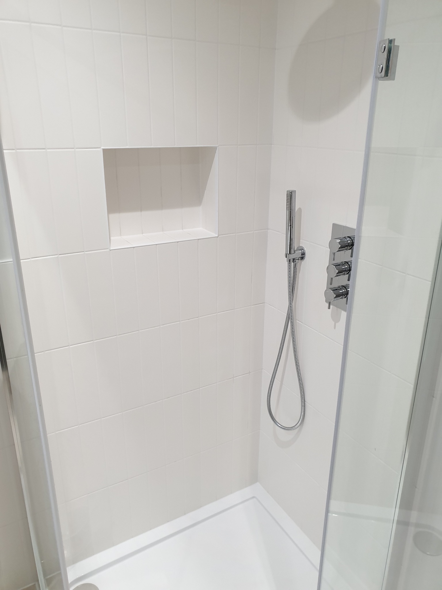 Nowoczesna kabina prysznicowa z białymi, prostokątnymi płytkami, chromowaną armaturą i wnęką w ścianie, widziana z perspektywy wnętrza kabiny.