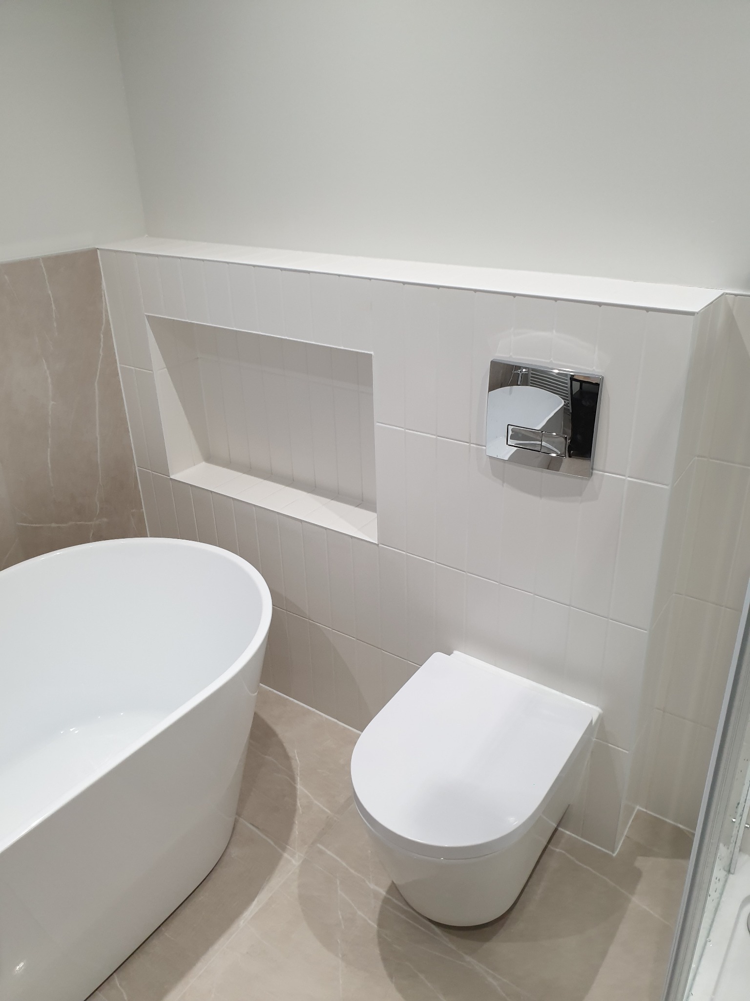 Nowoczesna łazienka z wolnostojącą wanną, podwieszanym WC z chromowanym przyciskiem spłukiwania i wnęką w ścianie wyłożonej białymi płytkami.