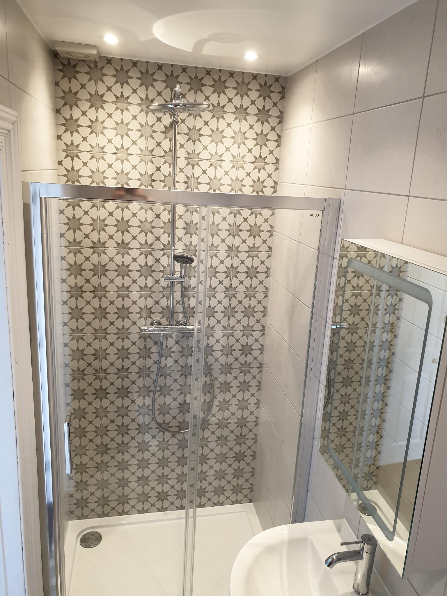 Wyremontowana łazienka z kabiną prysznicową ze srebrnymi profilami, wzorzystymi płytkami w odcieniach szarości na ścianie prysznica oraz białą umywalką z lustrem.