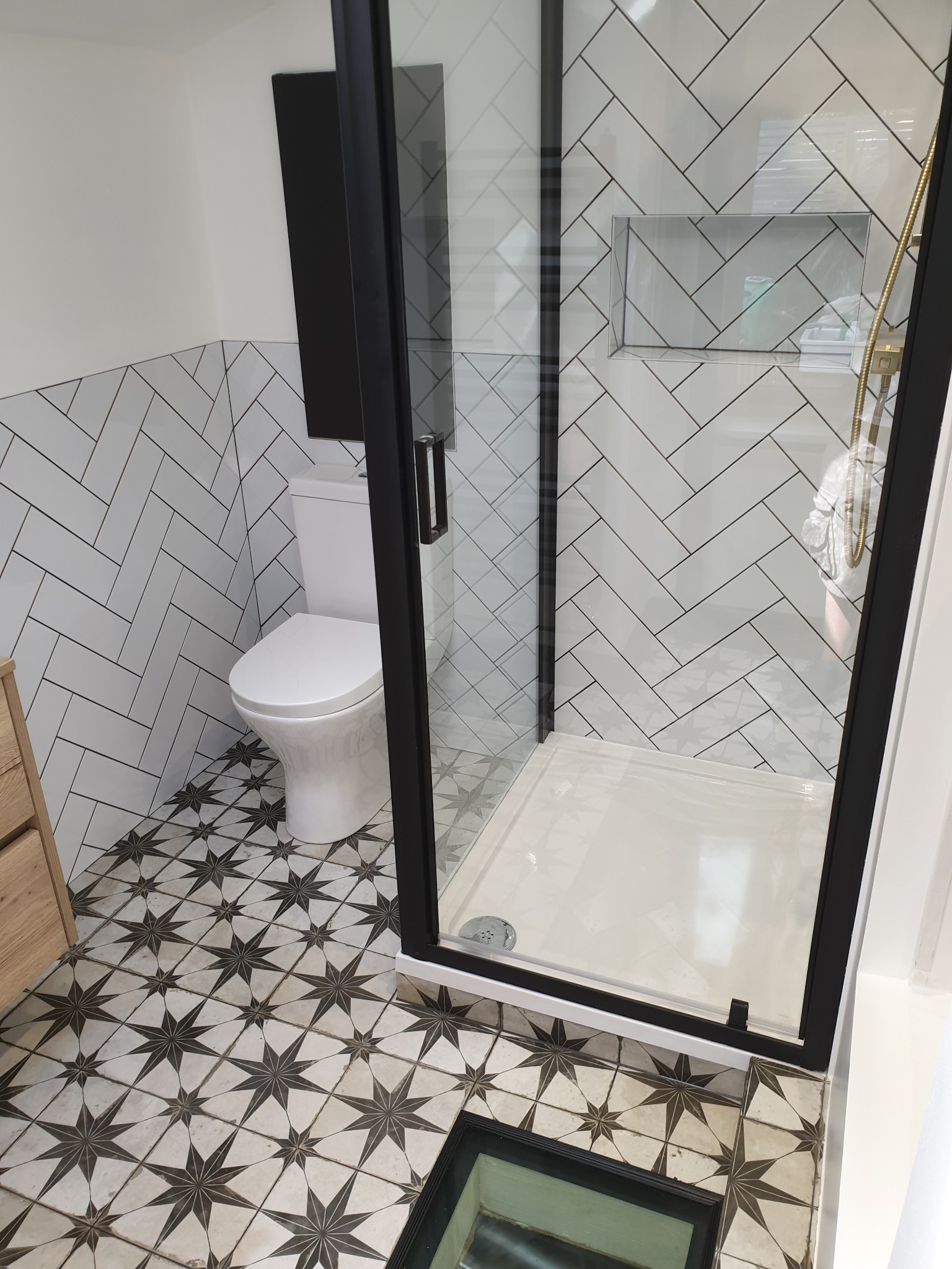 Nowoczesna łazienka po renowacji: kabina prysznicowa z czarną ramą, białe płytki ułożone w jodełkę, toaleta typu compact i podłoga w czarno-białe gwiazdy.