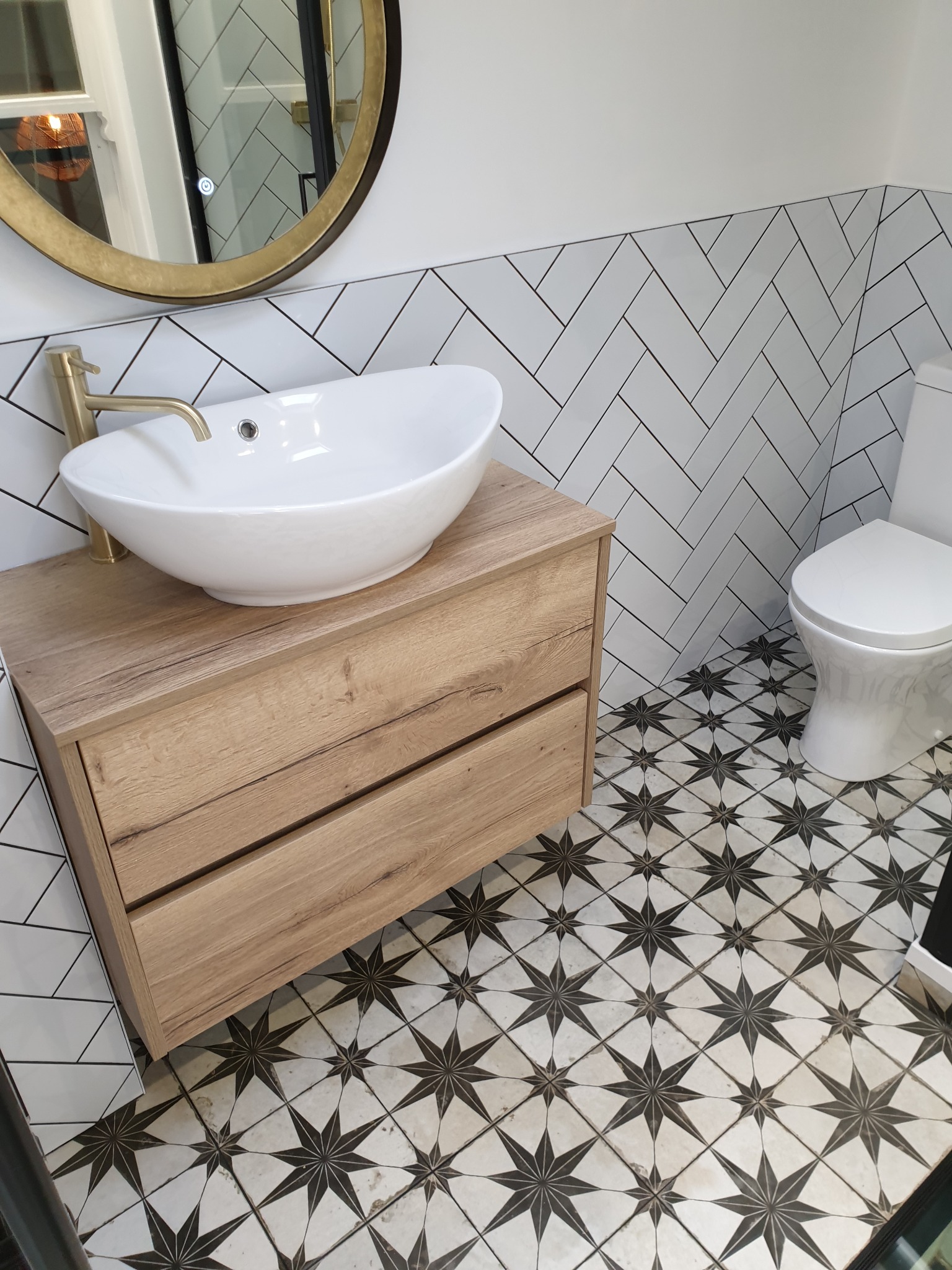 Nowoczesna łazienka z białą umywalką nablatową, złotą armaturą, szafką z drewnopodobnym dekorem, biało-czarnymi płytkami podłogowymi w gwiazdy i białymi płytkami ściennymi ułożonymi w jodełkę.