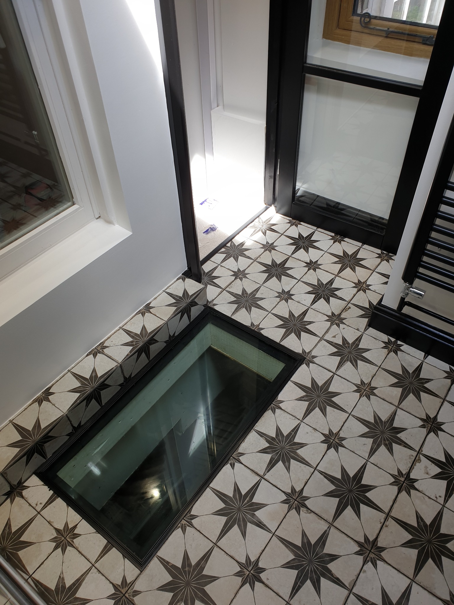 Podłoga wyłożona wzorzystymi płytkami z wbudowanym szklanym oknem, widok z góry, fragment grzejnika drabinkowego po prawej stronie.