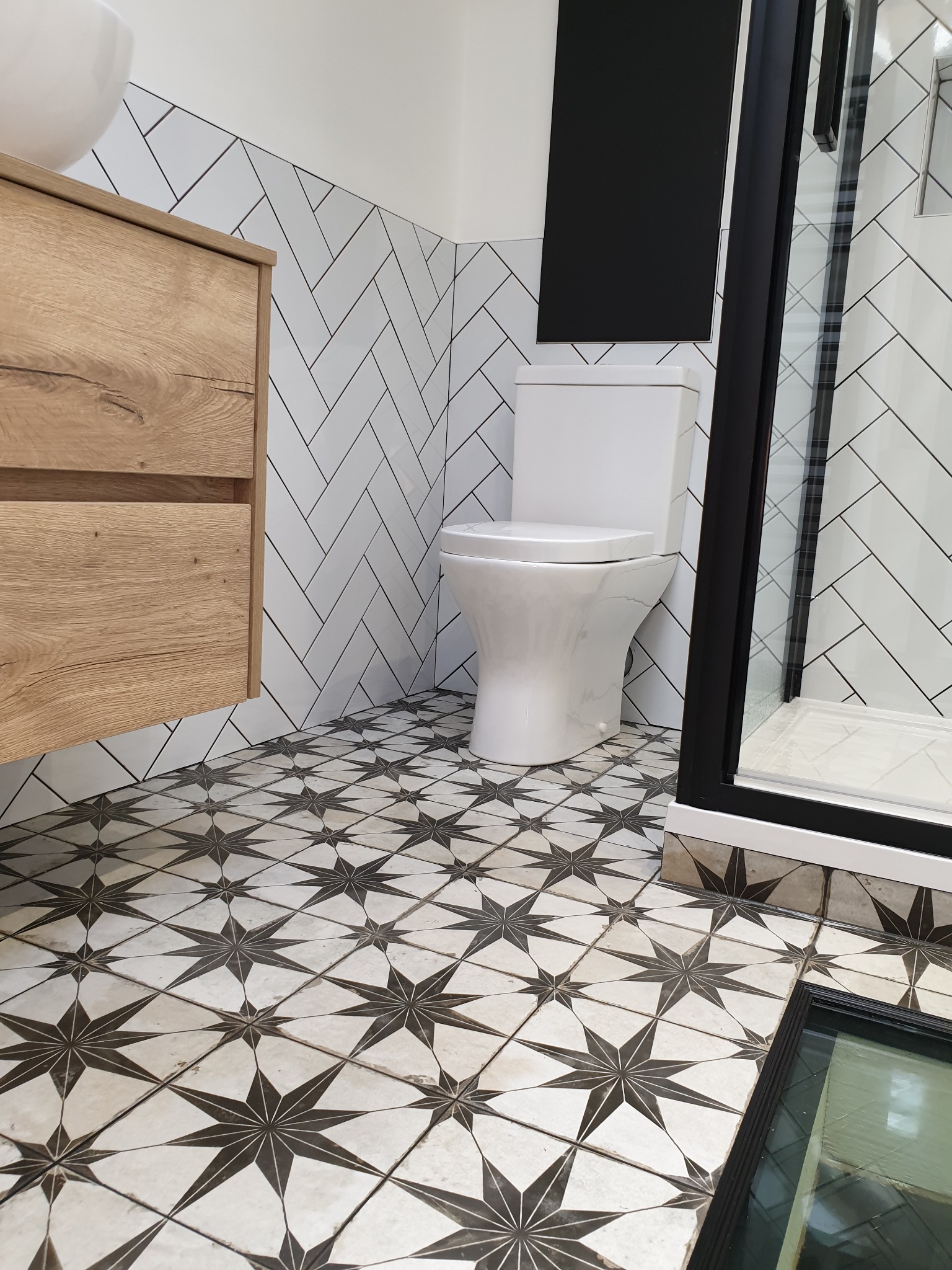 Nowoczesna łazienka z białą armaturą, drewnianą szafką pod umywalką i podłogą w czarno-białe gwiazdy. Ściany wyłożone białymi płytkami ułożonymi w jodełkę. Widoczna kabina prysznicowa z czarną ramą.