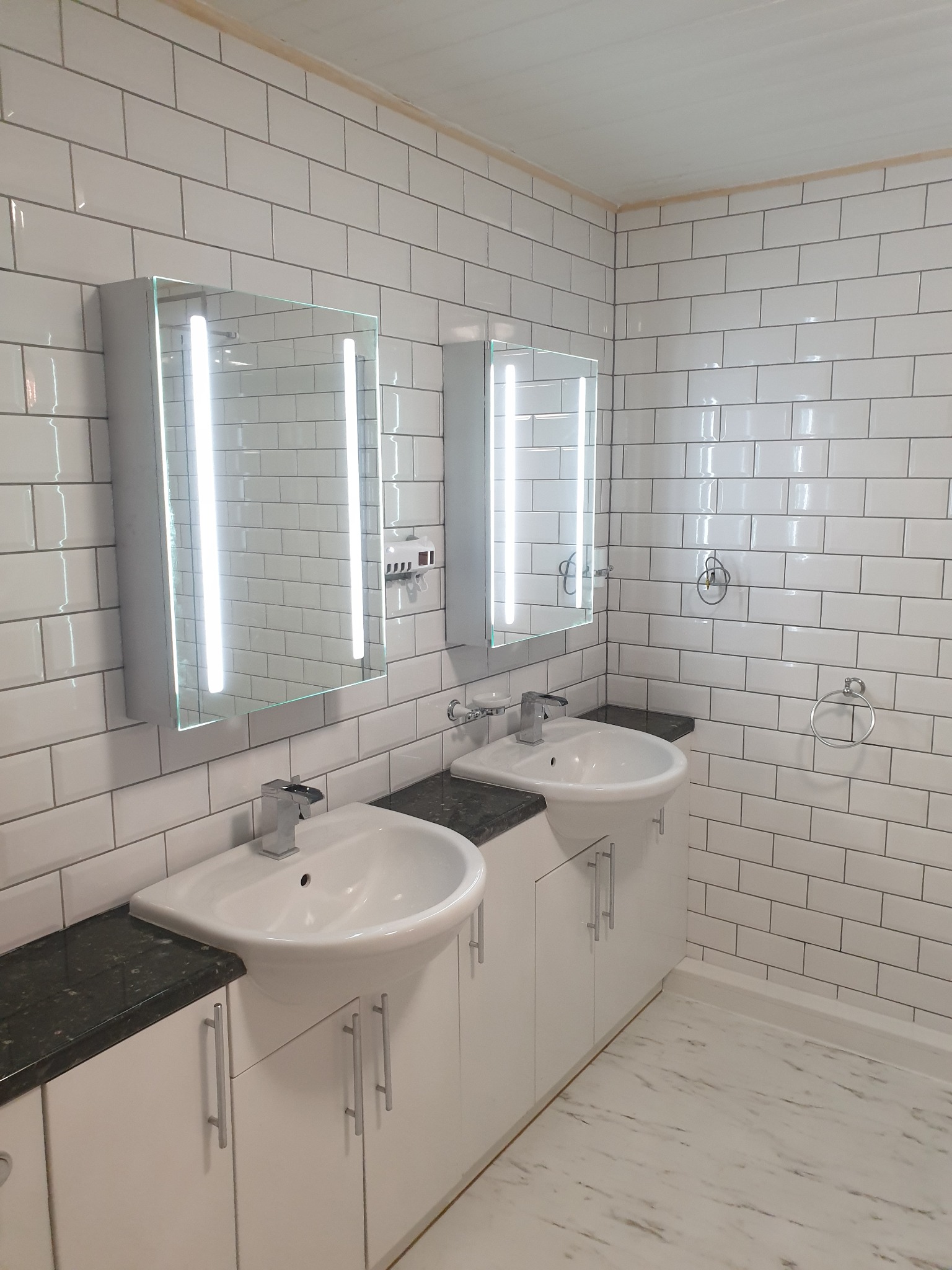 Nowoczesna łazienka z dwiema umywalkami nablatowymi na białych szafkach z ciemnymi blatami, podświetlane lustra, białe kafelki na ścianach i marmurowe panele na podłodze.