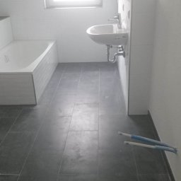 Mariusz Iwaszkiewicz - Wnętrze łazienki w trakcie prac wykończeniowych, widoczna wanna z obudową, umywalka oraz fragment podłogi z ciemnych płytek, wystające rury instalacyjne przy ścianie.