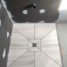 Ułożona podłoga w kabinie prysznicowej z jasnych, marmurkowych płytek z czarnymi fugami, widoczny odpływ liniowy, ściany w trakcie przygotowania pod płytki.