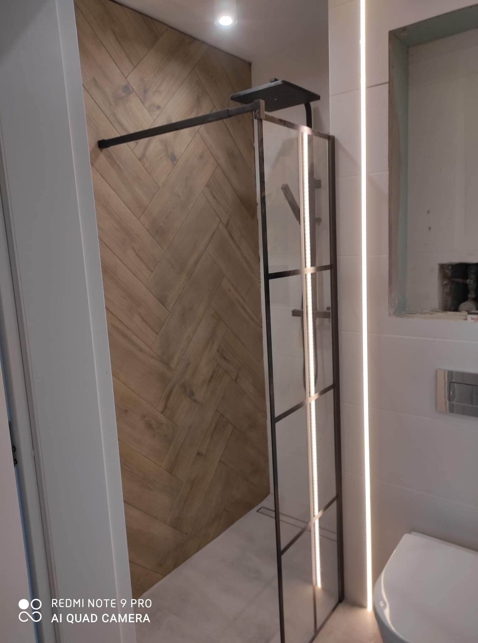 Nowoczesna kabina prysznicowa z czarną ramą i deszczownicą, zamontowana w łazience z drewnopodobnymi płytkami ułożonymi w jodełkę na jednej ścianie i białymi, podświetlanymi płytkami na drugiej...