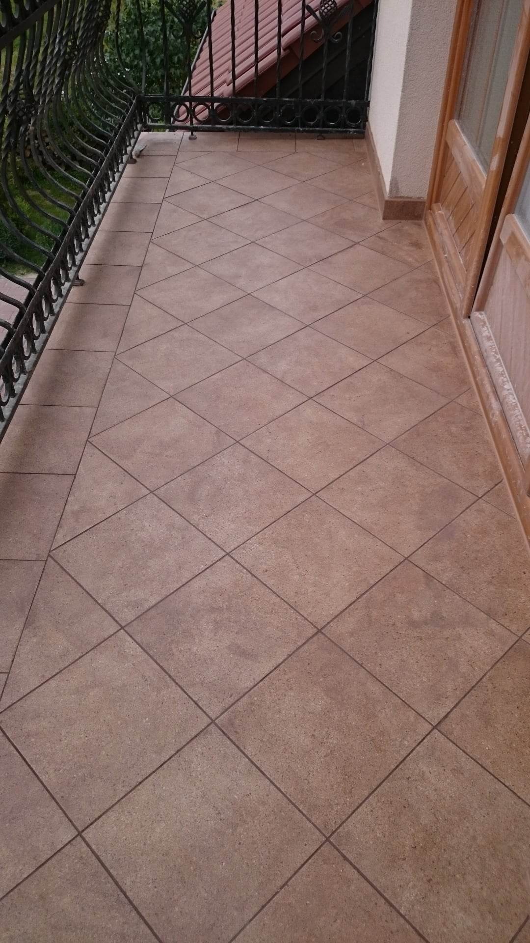 Wykończony balkon w Chełmie z brązowymi płytkami ułożonymi w romby, czarna, ozdobna balustrada i drewniane drzwi balkonowe.