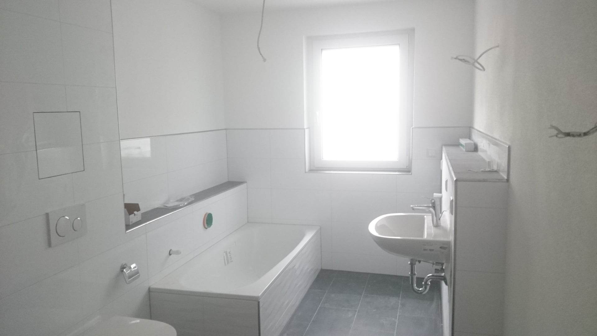 Jasna łazienka w trakcie wykańczania: białe płytki, wanna z obudową, umywalka z szafką i szare płytki podłogowe; widoczne wystające przewody elektryczne.