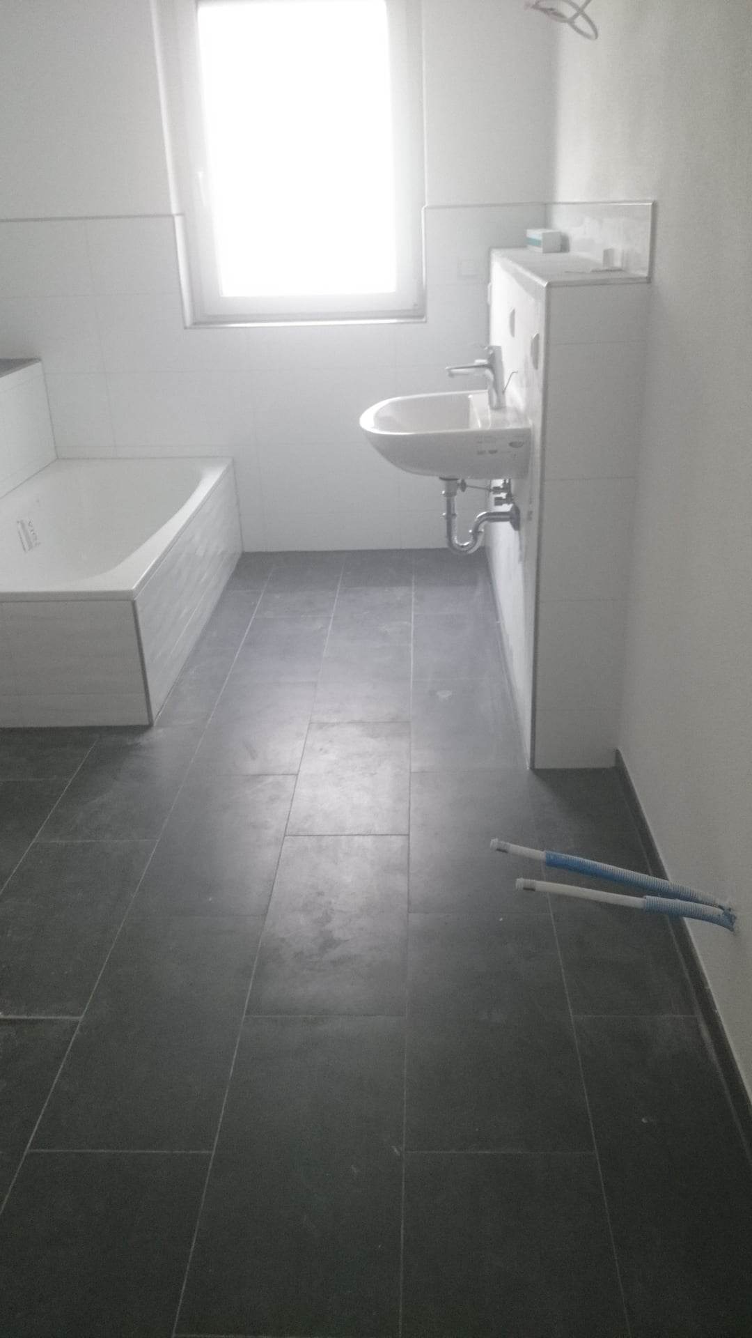 Wnętrze łazienki w trakcie prac wykończeniowych, widoczna wanna z obudową, umywalka oraz fragment podłogi z ciemnych płytek, wystające rury instalacyjne przy ścianie.