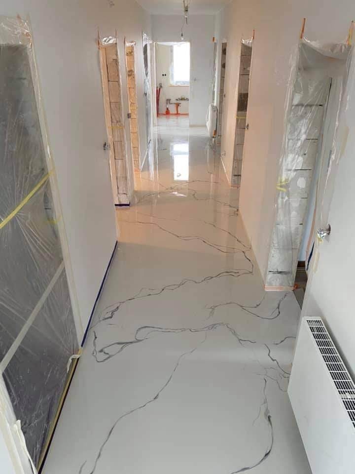 Korytarz w trakcie remontu z nową, błyszczącą podłogą imitującą marmur, zabezpieczone folią drzwi i grzejnik, widoczne narzędzia malarskie w oddali.