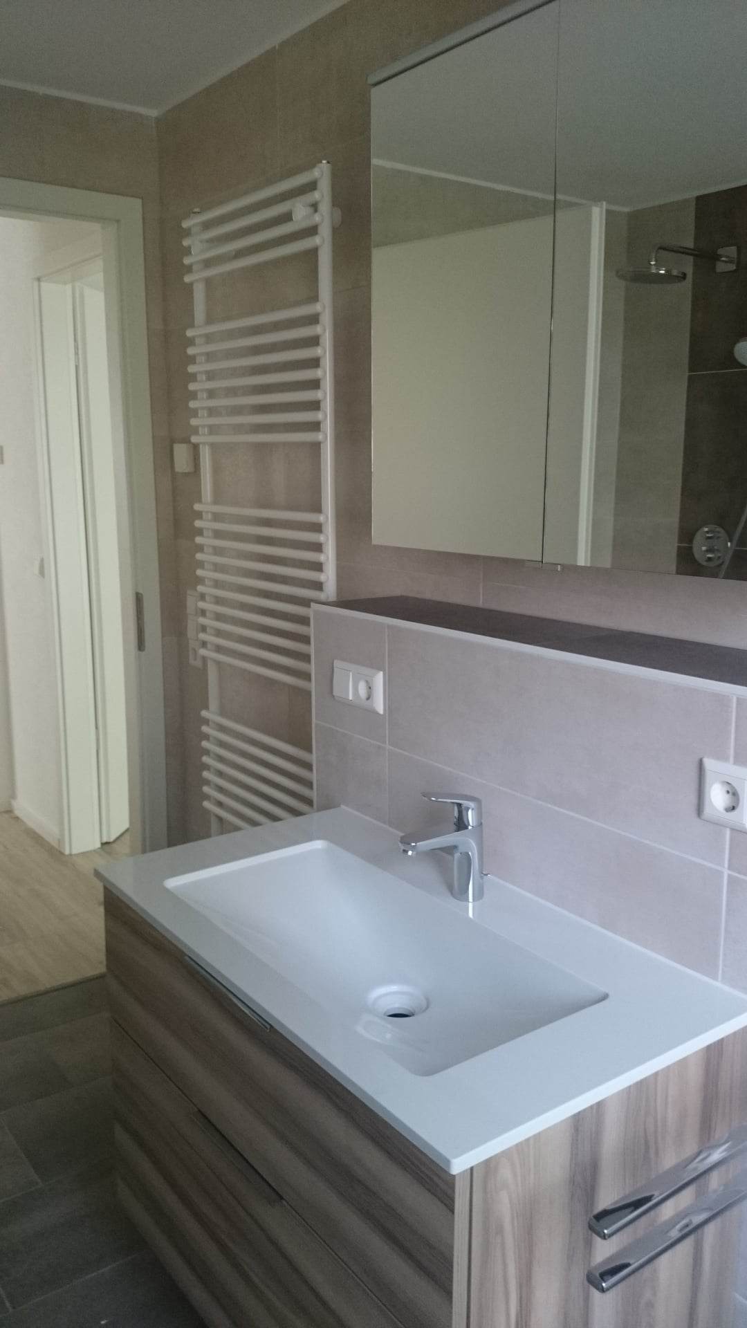 Nowoczesna łazienka z umywalką nablatową na szafce z drewnopodobnym fornirem, grzejnikiem drabinkowym i lustrem w zabudowie, widok na kabinę prysznicową w tle.