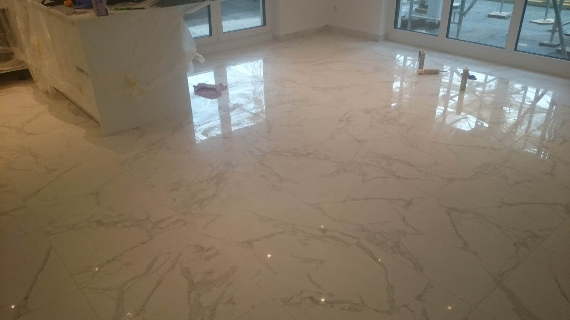 Świeżo położone, błyszczące, marmurowe płytki podłogowe w jasnym pomieszczeniu, z widocznymi materiałami budowlanymi przy oknie i zakrytą folią wyspą kuchenną.