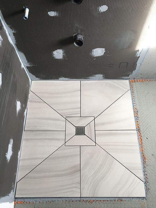 Ułożona podłoga w kabinie prysznicowej z jasnych, marmurkowych płytek z czarnymi fugami, widoczny odpływ liniowy, ściany w trakcie przygotowania pod płytki.