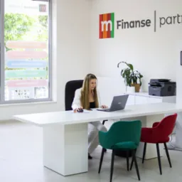 Kobieta przy biurku z laptopem w jasnym biurze z logo 'Finanse partner' i oknem z naklejkami informacyjnymi o kredytach.