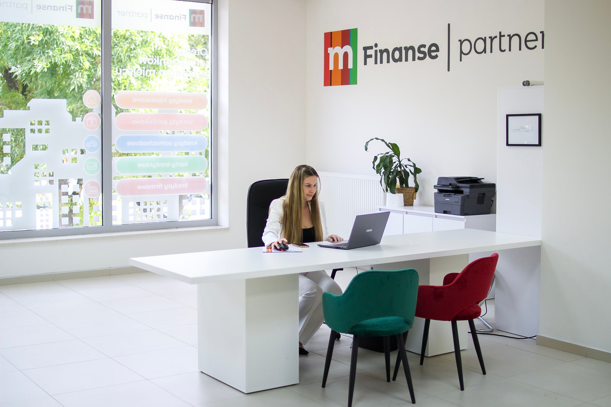 Kobieta przy biurku z laptopem w jasnym biurze z logo 'Finanse partner' i oknem z naklejkami informacyjnymi o kredytach.