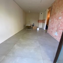 Vadym Bokhonovskyi West-Bud - Przestronne wnętrze w trakcie prac wykończeniowych: ułożone szare płytki podłogowe, ściany częściowo pomalowane na beżowo i różowo, wolnostojąca wanna na nóżkach w oddali, widoczne materiały...