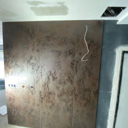 Vadym Bokhonovskyi West-Bud - Montaż dużych, brązowo-beżowych płyt imitujących rdzę na ścianie w trakcie prac wykończeniowych. Widoczne otwory na instalacje, przewody elektryczne oraz fragment sufitu z niezamaskowanymi kablami.