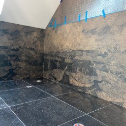 Vadym Bokhonovskyi West-Bud - Ściana łazienki z tapetą w stylu marynistycznym przedstawiającą żaglowce i miasto portowe, połączona z górną częścią ściany wykończoną metalową blachą ryflowaną, zabezpieczoną niebieską taśmą...