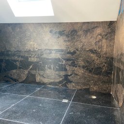 Vadym Bokhonovskyi West-Bud - Łazienka w trakcie prac wykończeniowych: czarna, błyszcząca podłoga z płytek, ściany pokryte tapetą z motywem historycznej bitwy morskiej, widoczny odpływ liniowy w podłodze i okno dachowe.